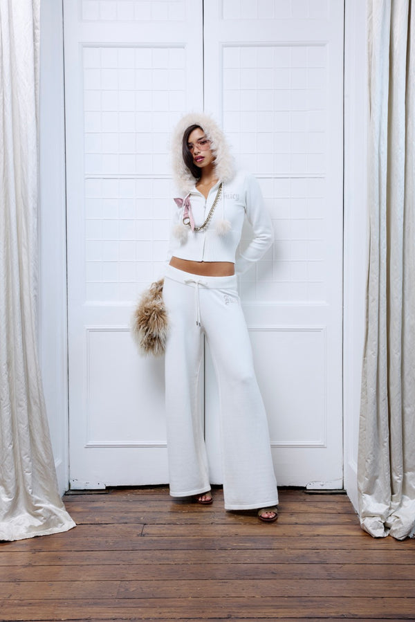 A woman stands in front of white paneled doors wearing an all-white outfit consisting of a cropped zip-up hoodie, a long white faux-fur hood, and matching wide-leg pants. She is holding a small pink bag and wearing a long gold chain necklace.
