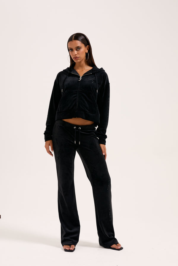 Displays a model wearing the Black outline 'Juicy' branded diamanté classic velour low rise pant 