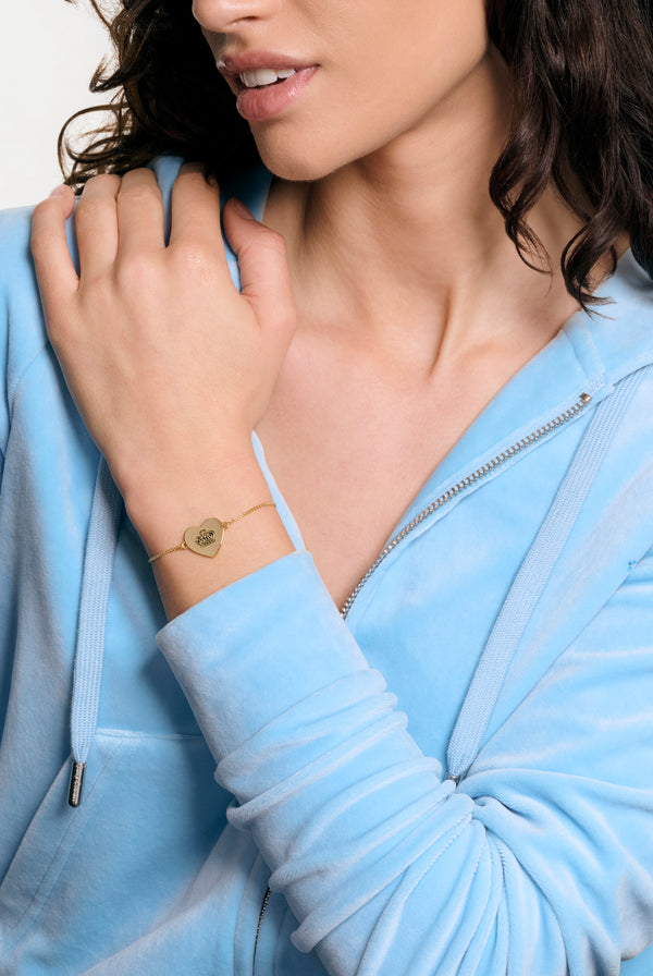 Displays a model wearing the gold tone 'Juicy' branded juicy Couture reversible heart adjustable bracelet 