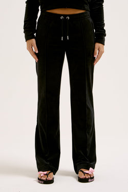 Woman wearing black velour tracksuit bottoms, with a black hoodie and black flip-flops on a white background
